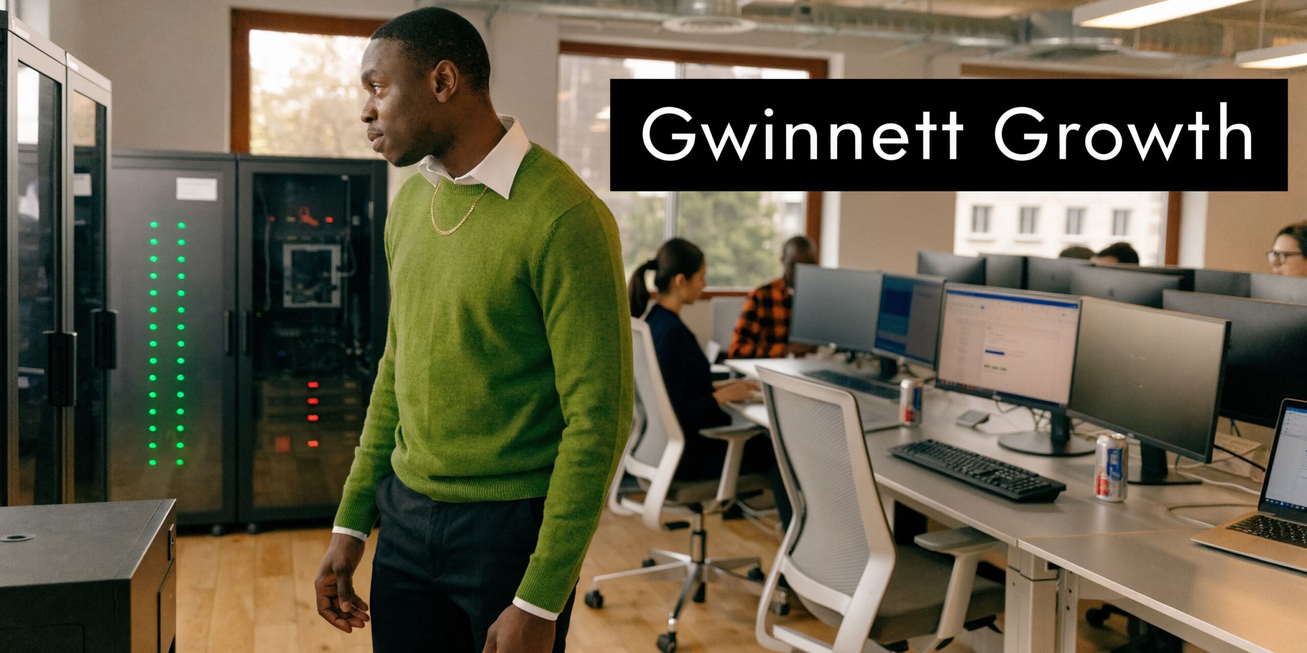 A professional man in a green sweater stands in a modern office with computer servers and workstations.